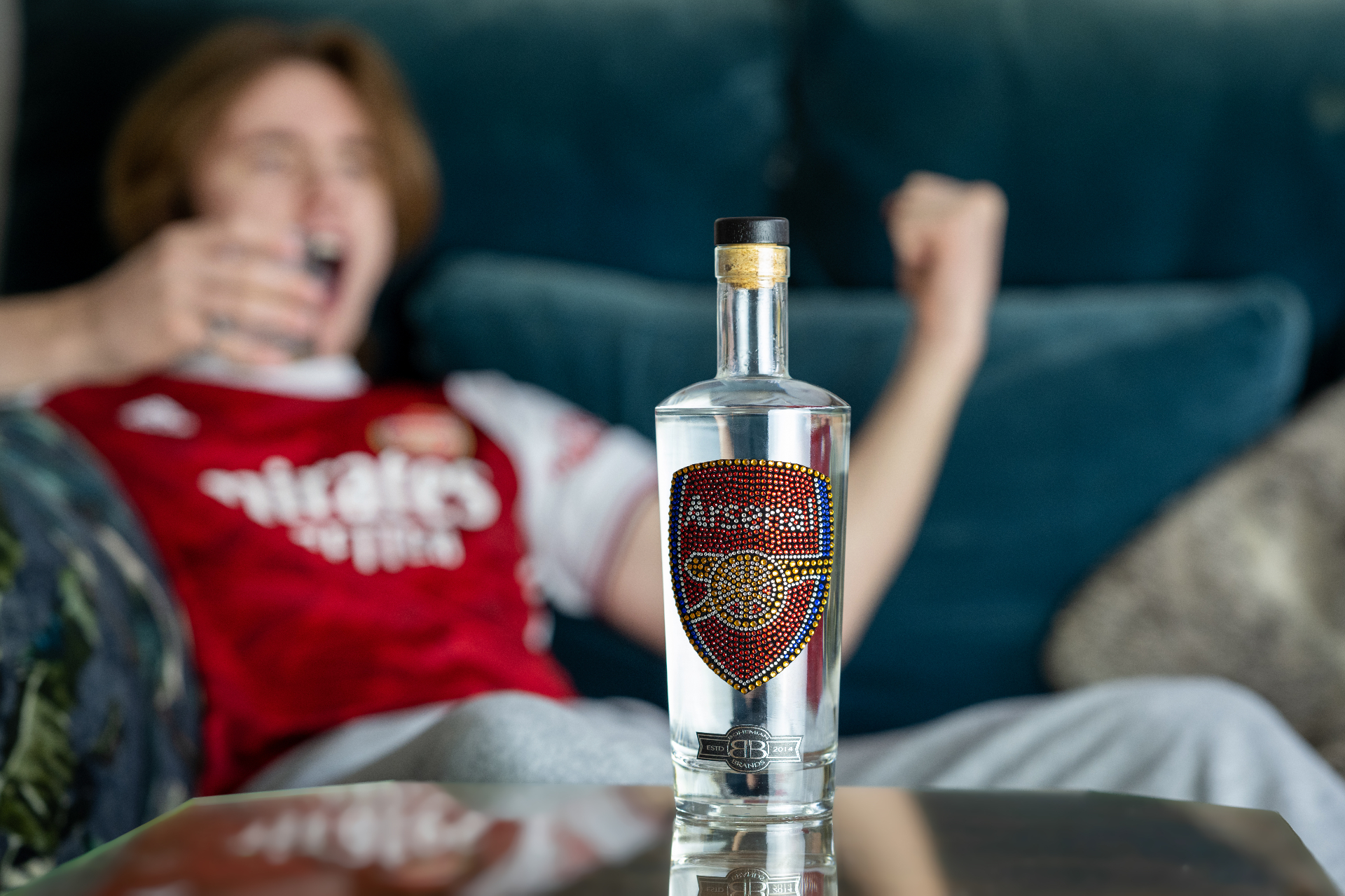 An Arsenal supporter celebrating on the sofa beside a Bohemian Brands crystal-crested bottle
